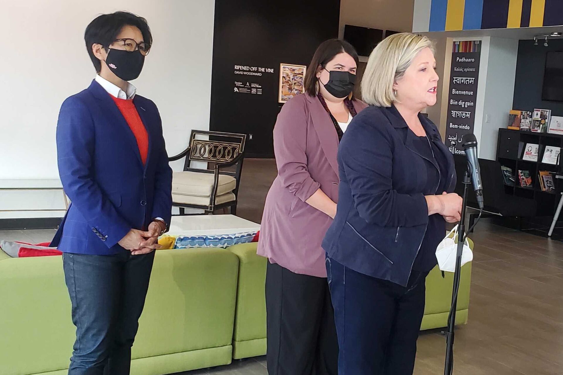 Three people standing in front of a green couch as they speak to a microphone. One person has a blue blazer, the person in the middle has a lavender coat and the person on the far right wears a navy jackey and has blonde hair.