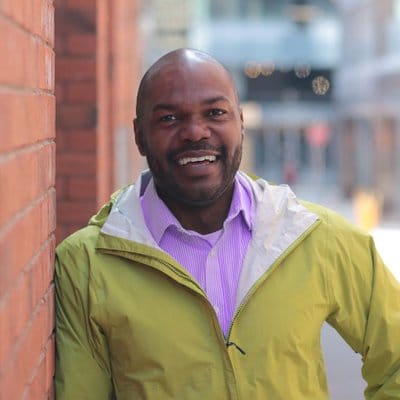 A man wearing a lavender dress shirt with a mustard jacket on top. He is smiling and leaning on a brick wall.