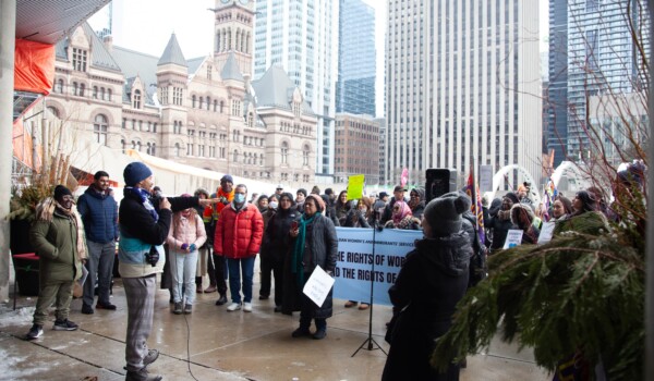 A crowd of people holding banners and signs in front of city hall. A speaker using a mic faces the ralliers.
