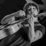 A black and white image of Alanna Matty strumming her acoustic guitar. She is wearing a short, flowy black dress and has tattoos on her arm. She has long brown hair up in a bun with bangs.