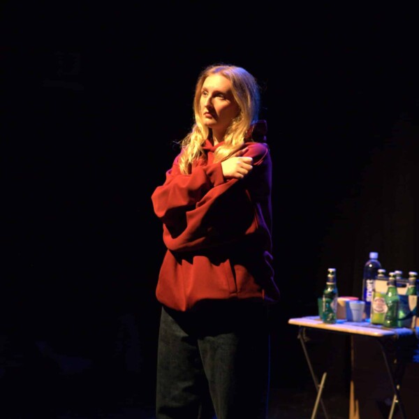 Actress dressed in black jeans, a red hoodie, with a concerned or somewhat exasperated look on her face, in a self hug sort of posture. She is lit by stage light, standing next to a small folding table with 4 small multicolored plastic tumbler glasses, a 6-pack of Peroni beer bottles, and a water bottle. The background is dark, making her a strong focal point amidst the black.