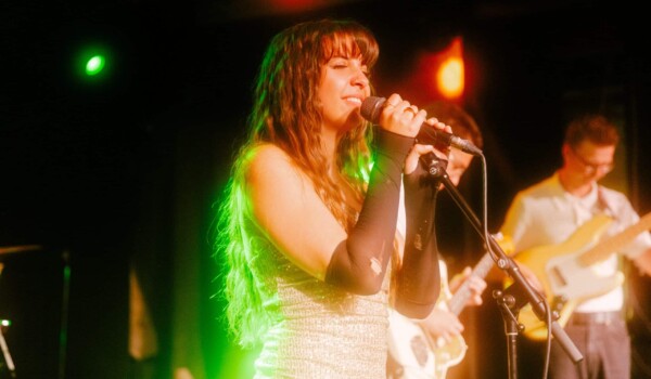 Ella Forrest, in a shimmery silver outfit, with long black fingerless gloves, singing on stage while holding a microphone resting in a stand. A stagelight behind her is illuminating her hair a bright green.