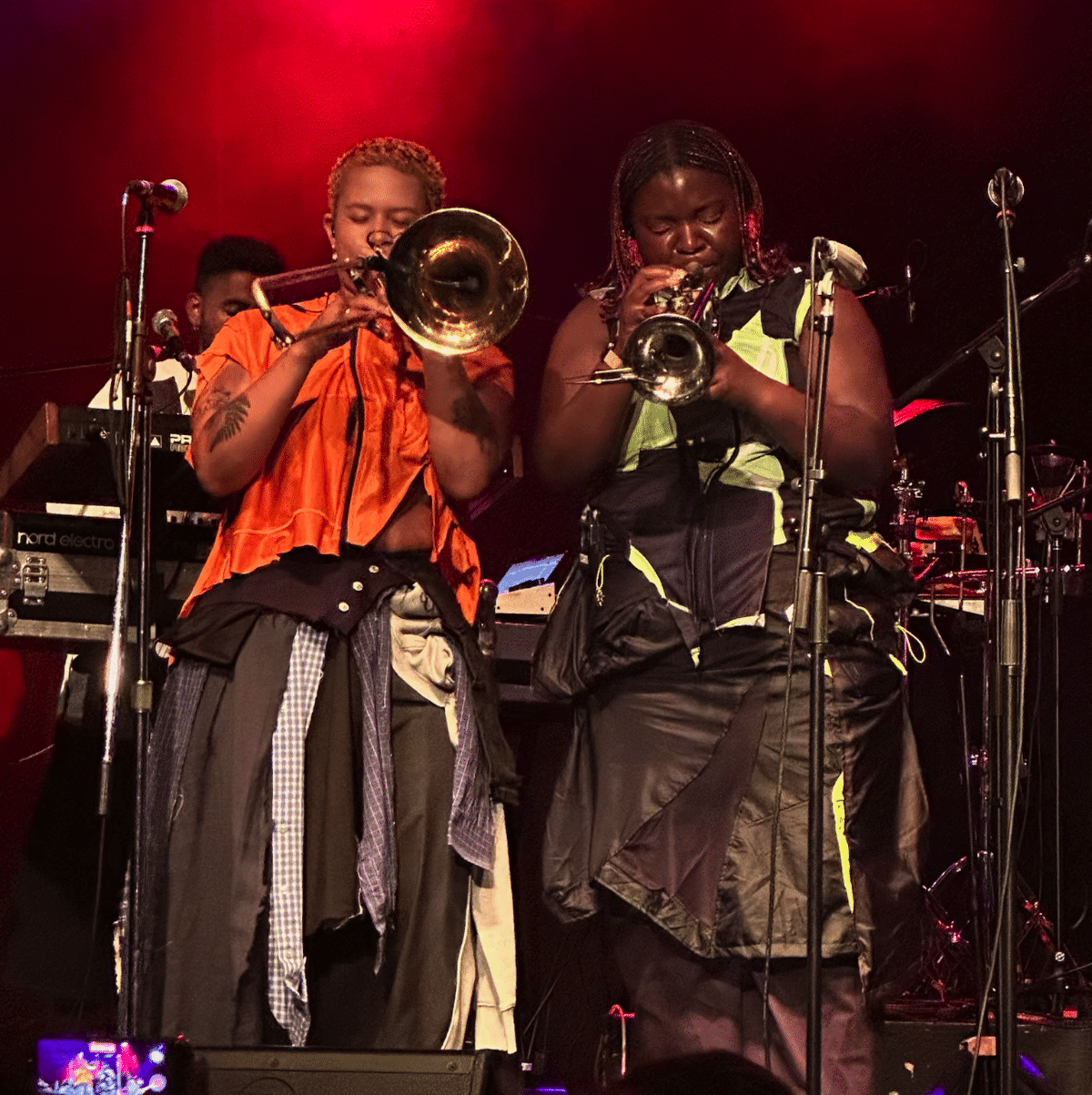 Two brass players of Kokoroko centre stage in bulky tattered, but collage-style fashion, one playing trombone and one trumpet, both with eyes closed in concentration, the keyboard player slightly visible in the background, all in front of a reddish hue from stage lights.