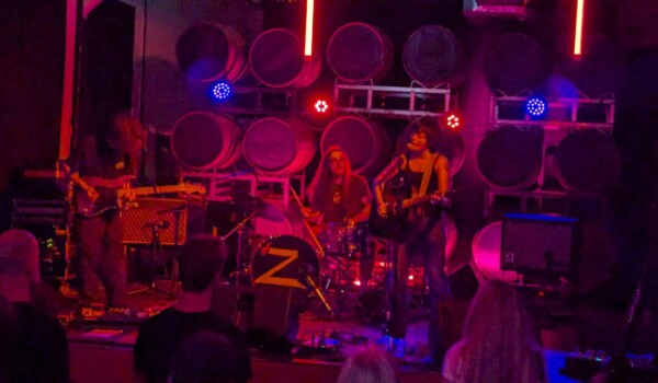 Zoh Amba in bluejeans and a black tank top plays an acoustic guitar, mouth open as she sings into a mic, with electric guitarist hanging his head down and his shaggy hair covering his face. A drummer in the background with long hair and square framed glasses plays along. The stage is in blue and red lights with the round tops of beer kegs lined in rows behind them as the stage backdrop.