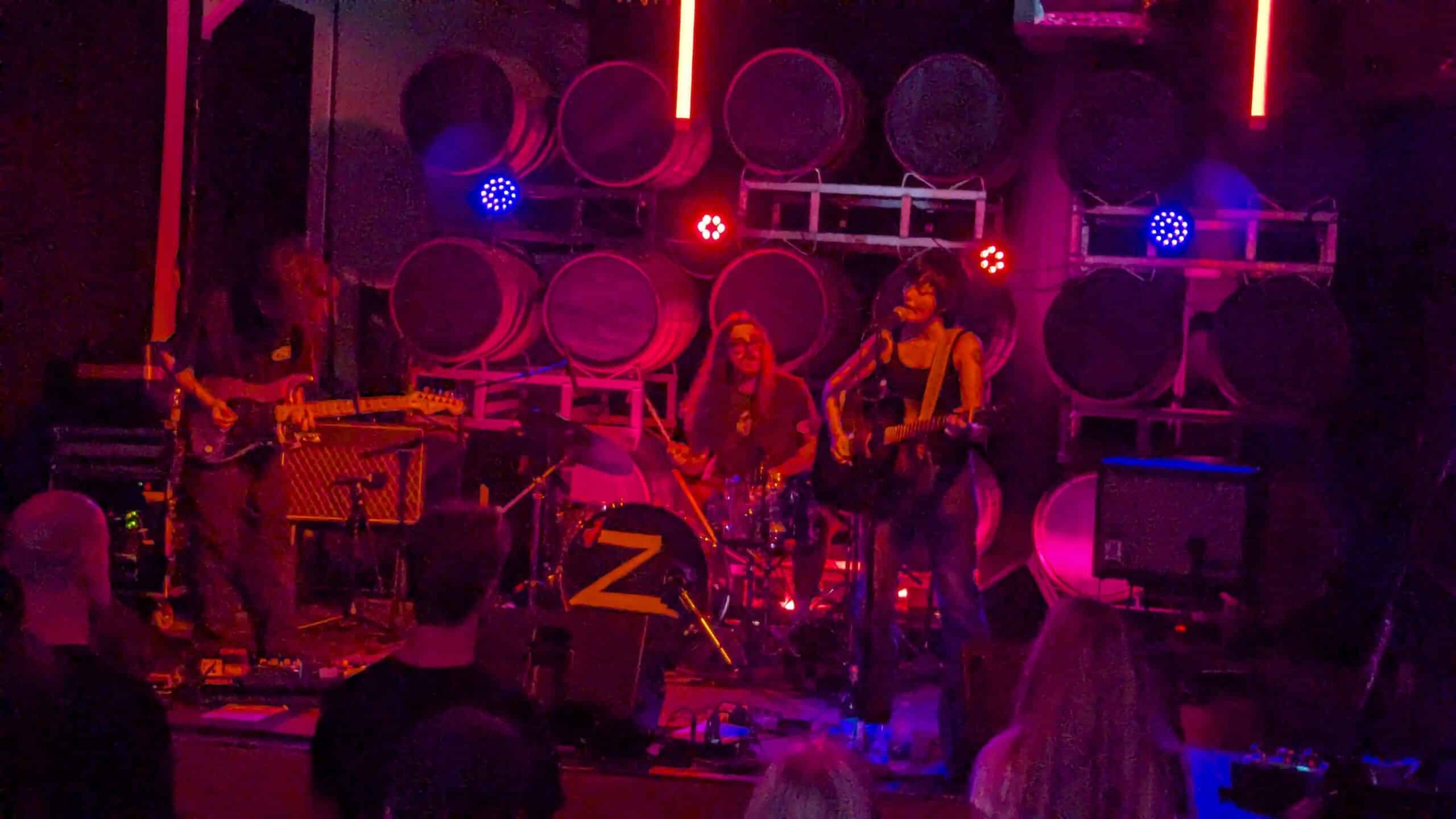 Zoh Amba in bluejeans and a black tank top plays an acoustic guitar, mouth open as she sings into a mic, with electric guitarist hanging his head down and his shaggy hair covering his face. A drummer in the background with long hair and square framed glasses plays along. The stage is in blue and red lights with the round tops of beer kegs lined in rows behind them as the stage backdrop.